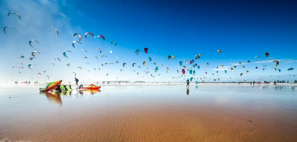 Kitesurfing, kite spot, wing in foil, množica kitov nad mokro peščeno plažo pod modrim nebom